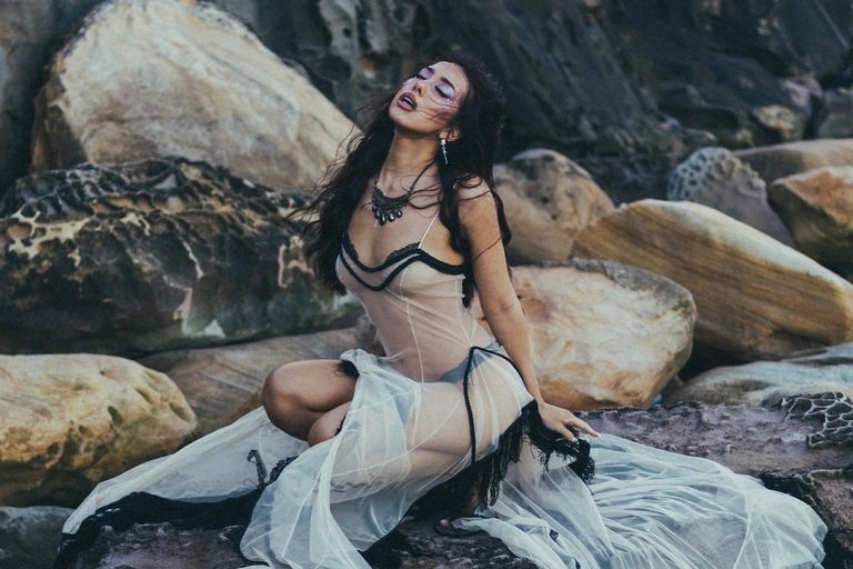 Musical artist Liliana de la Rosa posing in a flowing sheer dress on rocky coastal terrain.